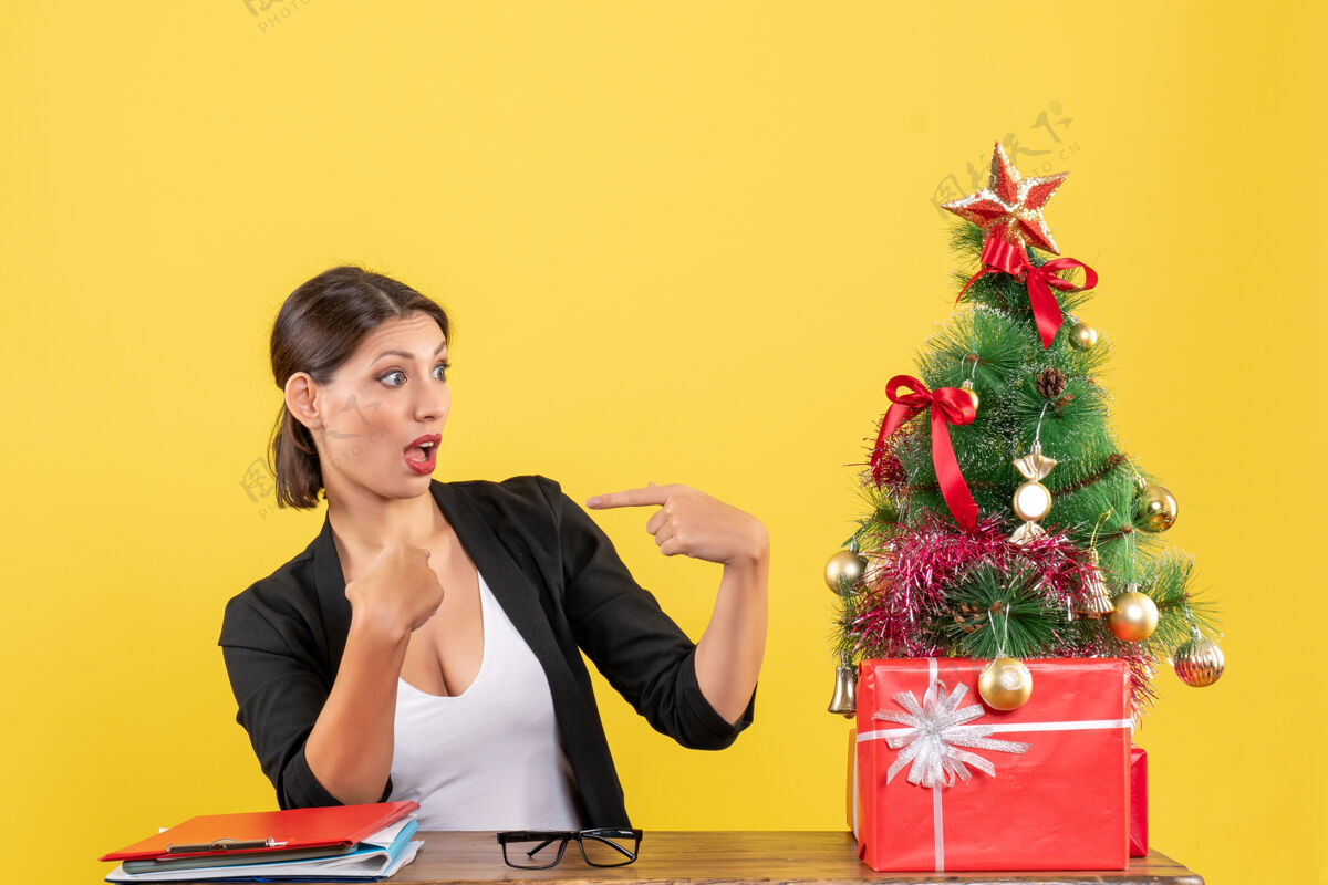 电脑一个穿着西装的年轻女子指着黄色的办公室里装饰好的圣诞树 惊讶不已坐着套装新郎