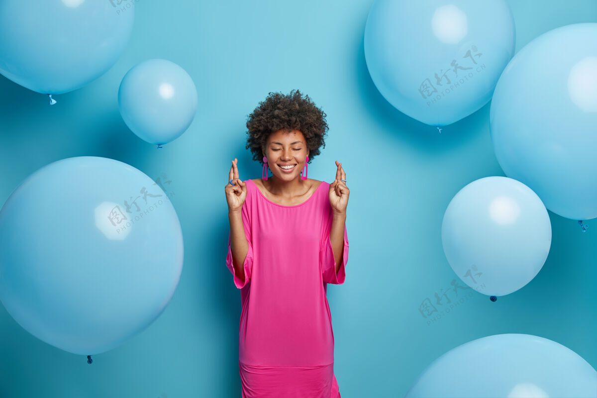手势积极的非裔美国妇女穿着优雅的粉色连衣裙 交叉手指 期待着一些令人向往的事情发生 在派对上 对着蓝色的墙壁摆姿势 周围放着充气的气球祈祷气球手指