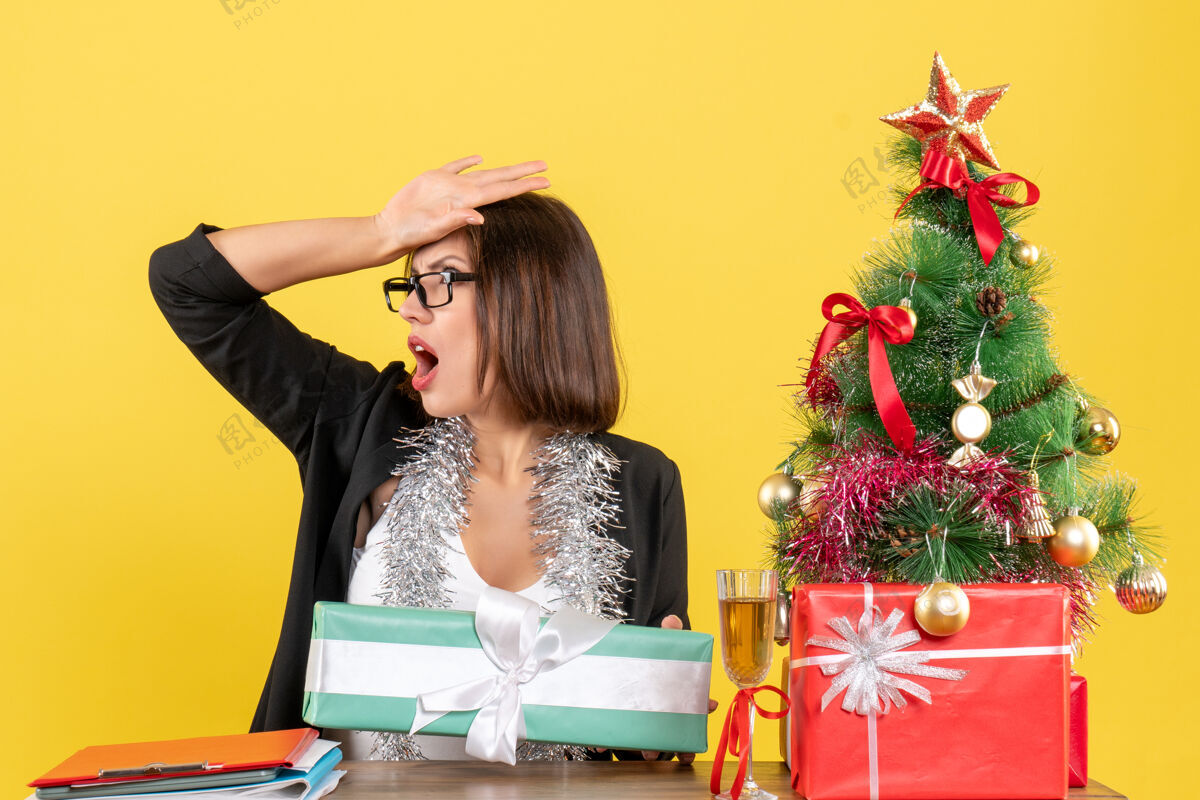 快乐一位穿着西装 戴着眼镜展示礼物 坐在办公室里一棵圣诞树旁的商务女士大吃一惊封面女郎树性感