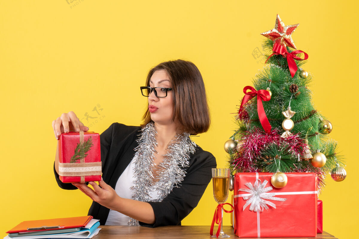 漂亮一位穿着西装 戴着眼镜的好奇的商务女士举起礼物 坐在办公室的一张桌子旁 桌上放着一棵圣诞树西装快乐高兴