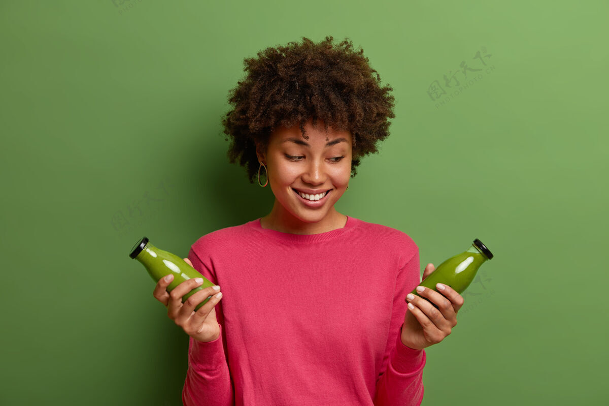 卷曲积极微笑的卷发女人摆出两瓶绿色排毒冰沙 喝健康新鲜的含维生素的素食饮料 穿粉色套头衫 坚持节食 在室内摆姿势素食生物心情