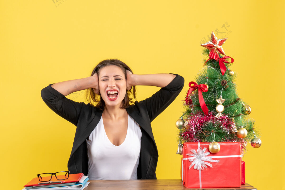 黄色情绪激动的快乐年轻女子闭上耳朵坐在黄色办公室装饰圣诞树旁的桌子旁模特人情感