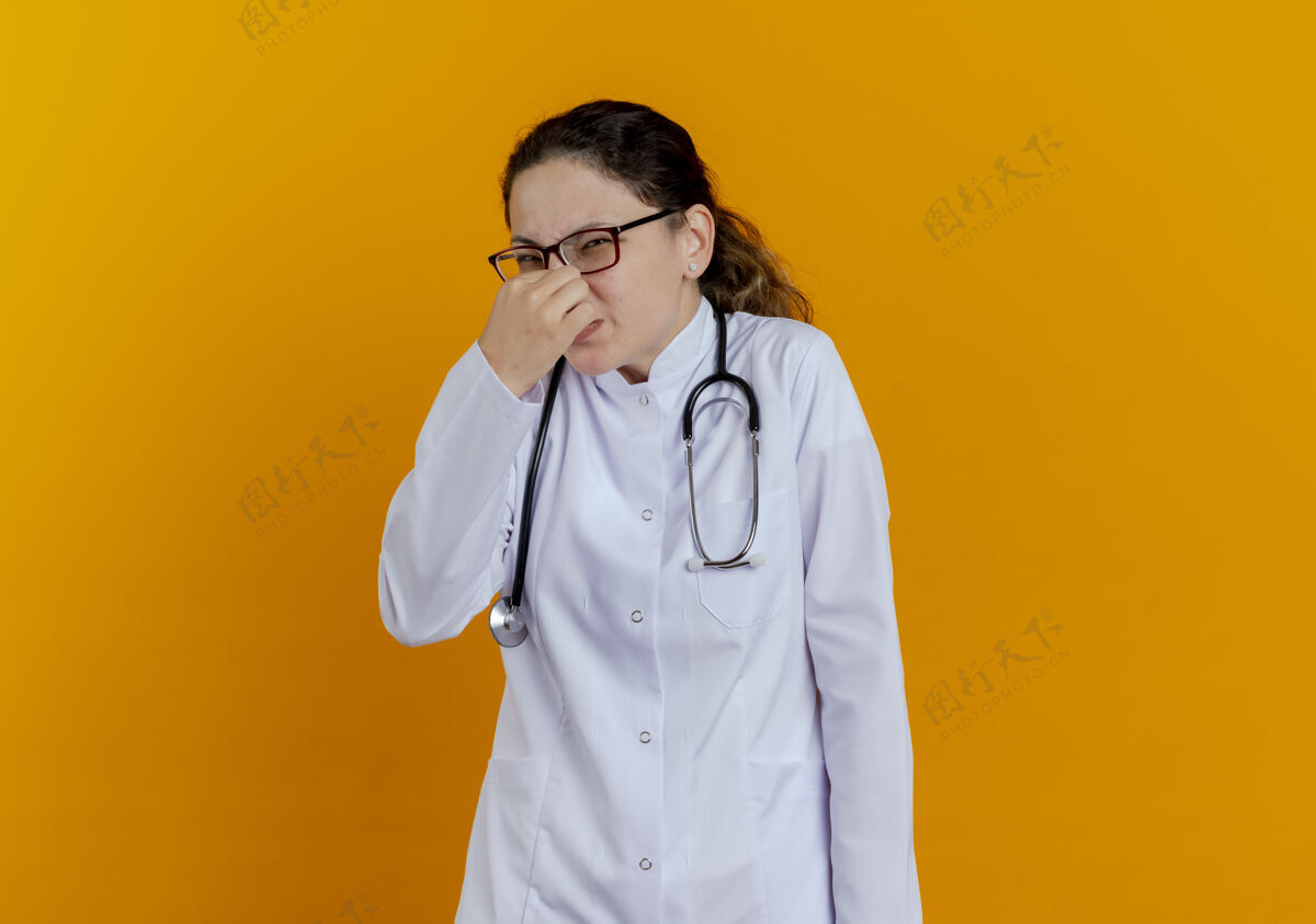 听诊器神经质的年轻女医生 穿着医用长袍 戴着听诊器 戴着眼镜 闭着鼻子年轻长袍成人