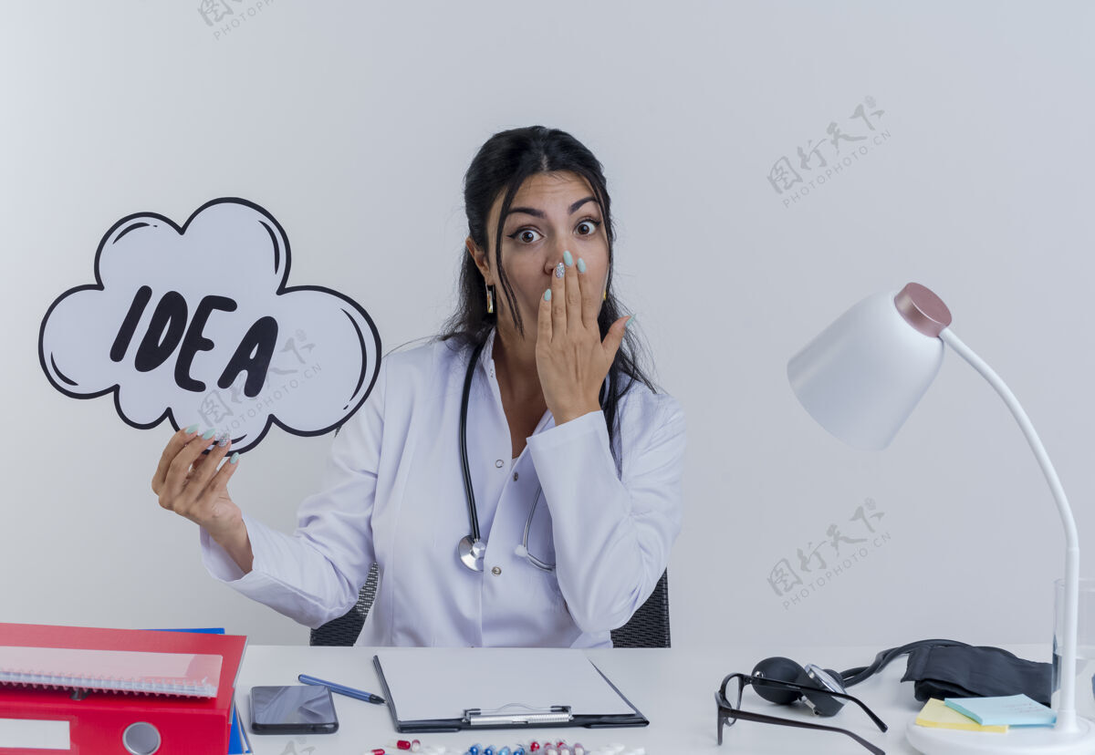 年轻人惊讶的年轻女医生穿着医用长袍和听诊器坐在办公桌旁 手里拿着医疗工具 手里拿着创意泡泡 看着把手放在嘴上人成人医学