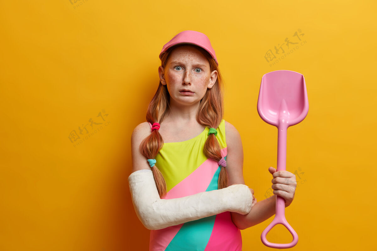 阴沉困惑的小女孩戴着帽子 穿着泳衣 拿着玩具沙铲 在海边的沙滩上玩耍 严重受伤后手臂缠着绷带 隔离在黄色的墙上童年和夏天的休息理念年轻外表受伤