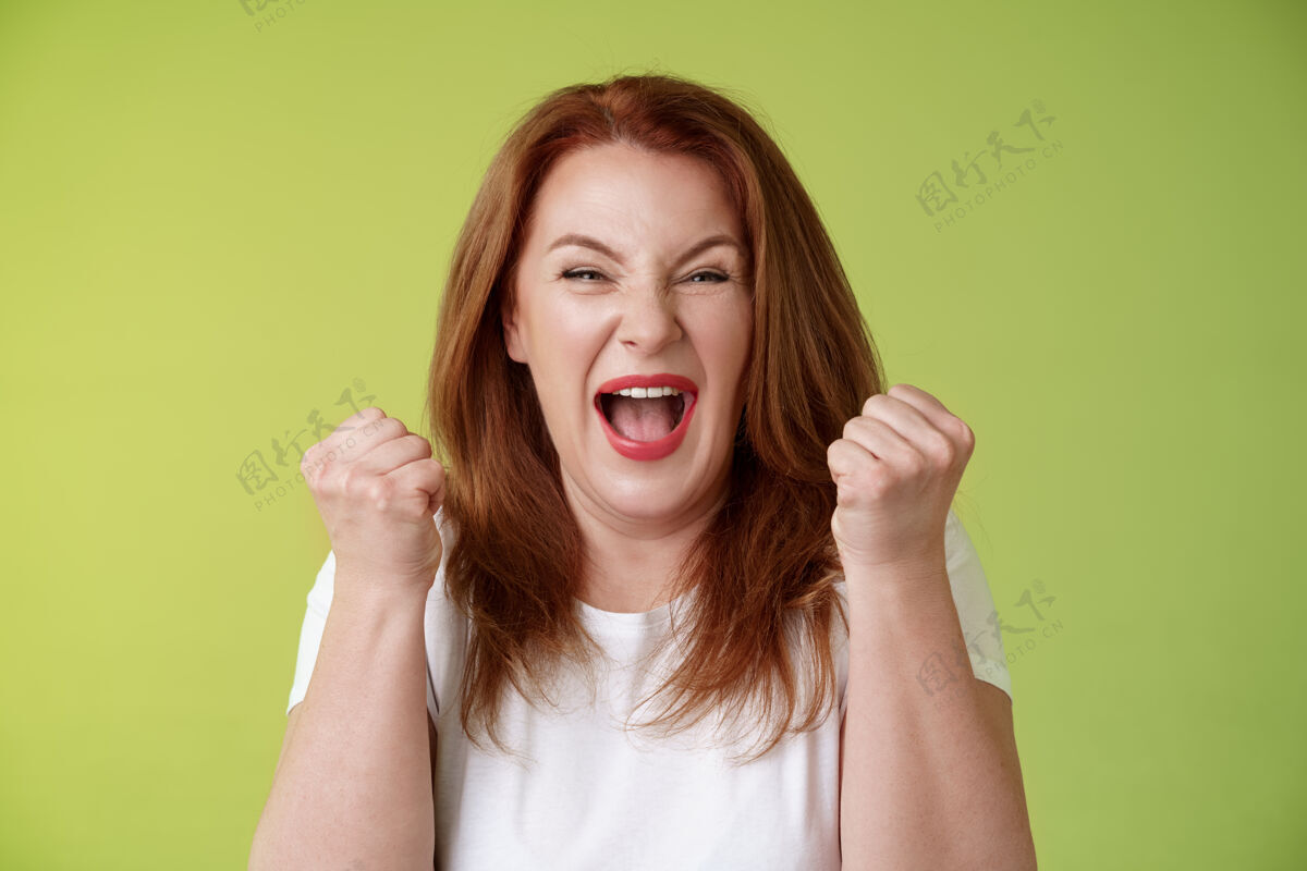 胜利是的 我们做到了快乐幸运的红发中年女性获胜者举起拳头庆祝成功手势大喊胜利快乐微笑广泛庆祝真棒的新闻站绿色墙快乐女性女人是