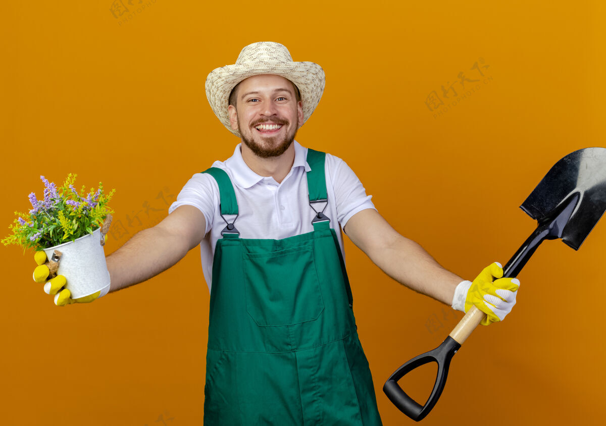 男人快乐的年轻英俊的斯拉夫园丁穿着制服戴着帽子和园艺手套伸出铁锹和花盆隔离家伙橙色手套