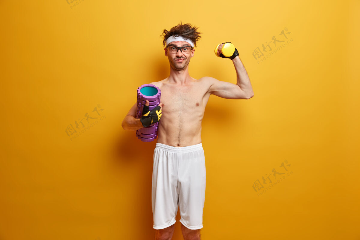 活动图为坚定的运动员举起哑铃 摆出泡沫滚轴姿势 目标是强壮 喜欢在健身房锻炼 隔离在黄色的墙壁上人 健康和健身的概念健康的生活方式健美肌肉健身房