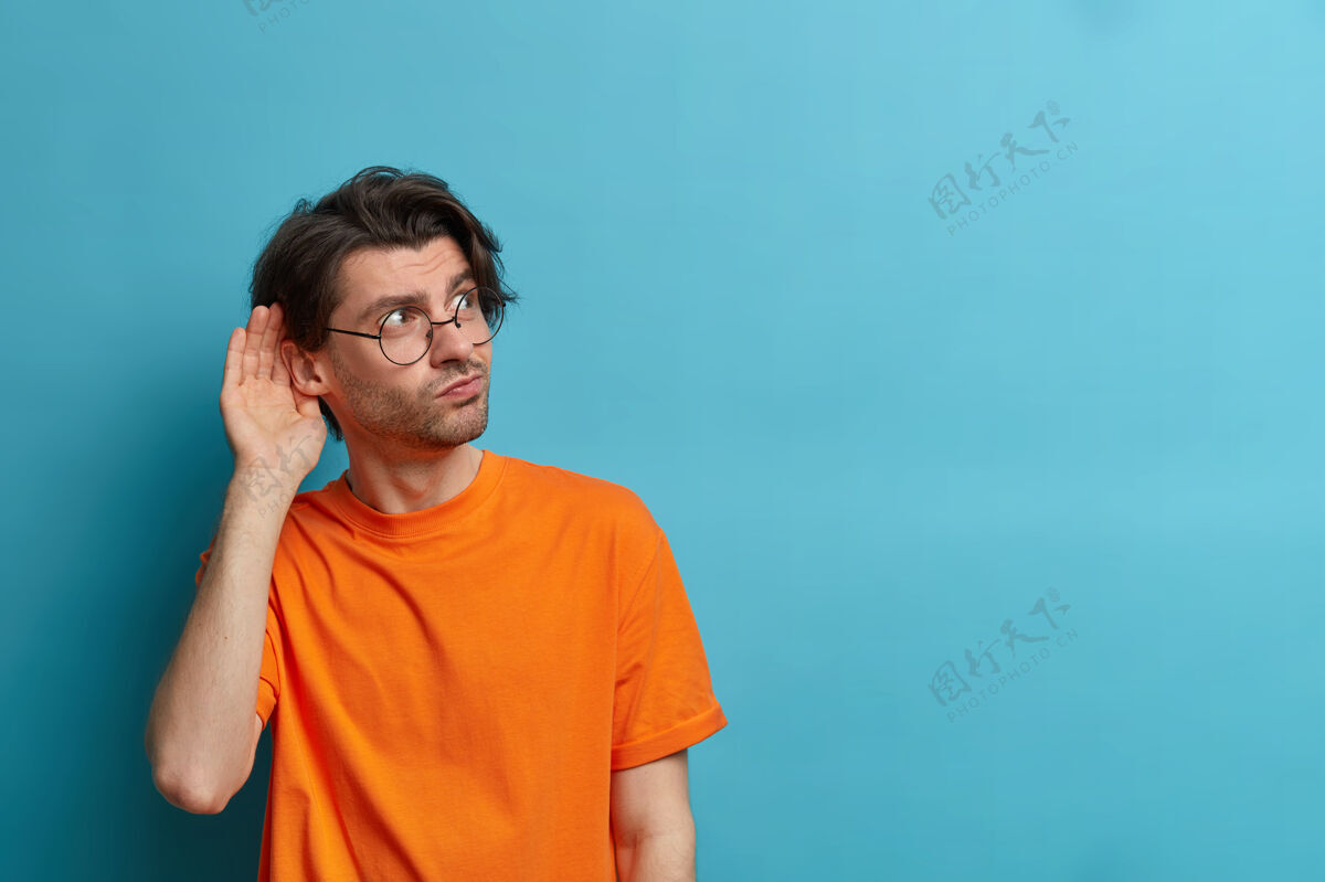 眼镜好奇男子的照片把手放在耳边 倾听私人信息 试图偷听闲话 有耐人寻味的表情 戴着圆眼镜和橙色t恤 在蓝色的墙上复制空间男人八卦秘密