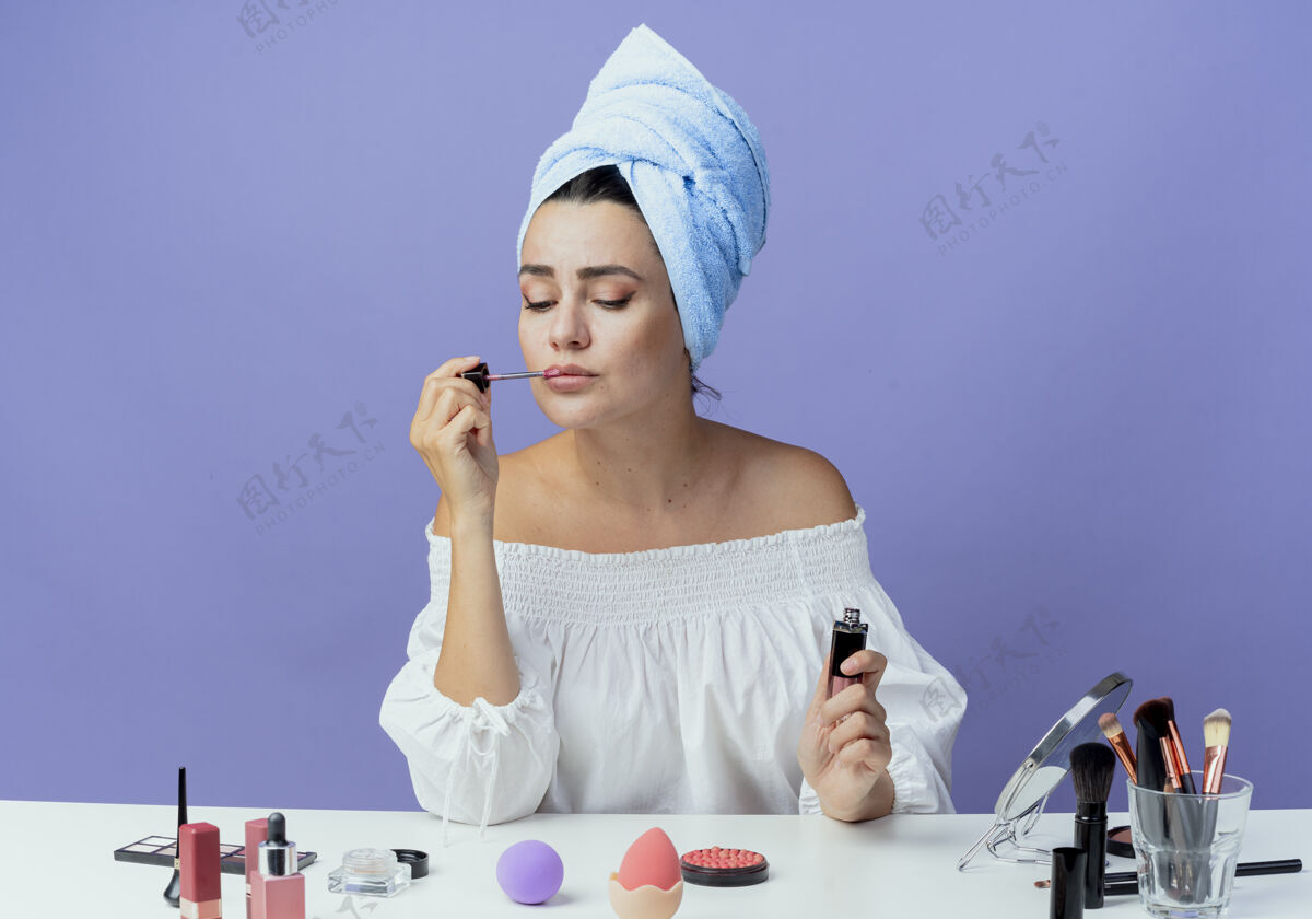 应用美丽的心烦意乱的女孩裹着发巾坐在桌子上 用化妆工具夹着唇彩 看着紫墙上的孤零零心烦嘴唇女人