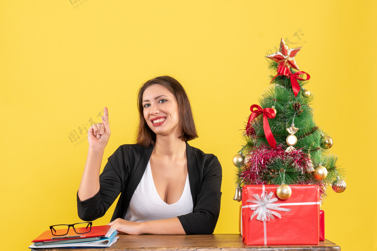 人一个年轻的女人坐在一张桌子旁 穿着西装指着黄色的办公室装饰的圣诞树指着套装快乐