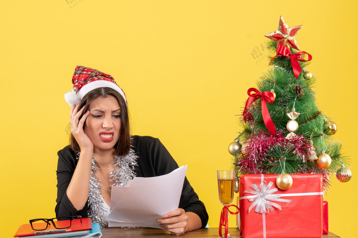 大人穿着西装 戴着圣诞老人帽 戴着新年装饰品的商务女士感觉筋疲力尽 坐在办公室的桌子旁 桌上放着圣诞树圣诞老人帽子西装