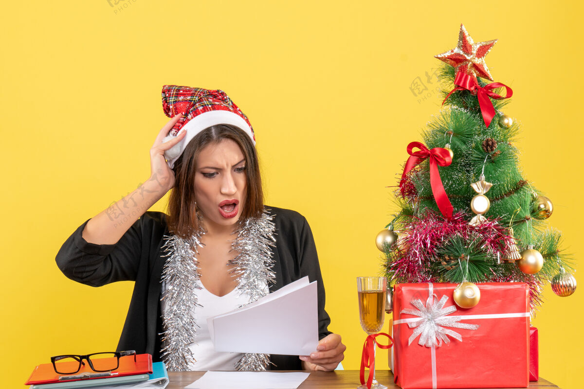 桌子穿着西装 戴着圣诞老人帽和新年装饰品的商务女士感到紧张 坐在办公室的桌子旁 桌上放着圣诞树帽子衣服感觉