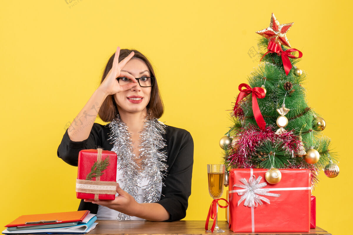 树一位身着西装 戴着眼镜的商务女士展示了她制作礼物的眼镜姿势 坐在办公室的一张桌子旁 桌上放着一棵圣诞树眼镜礼物展示