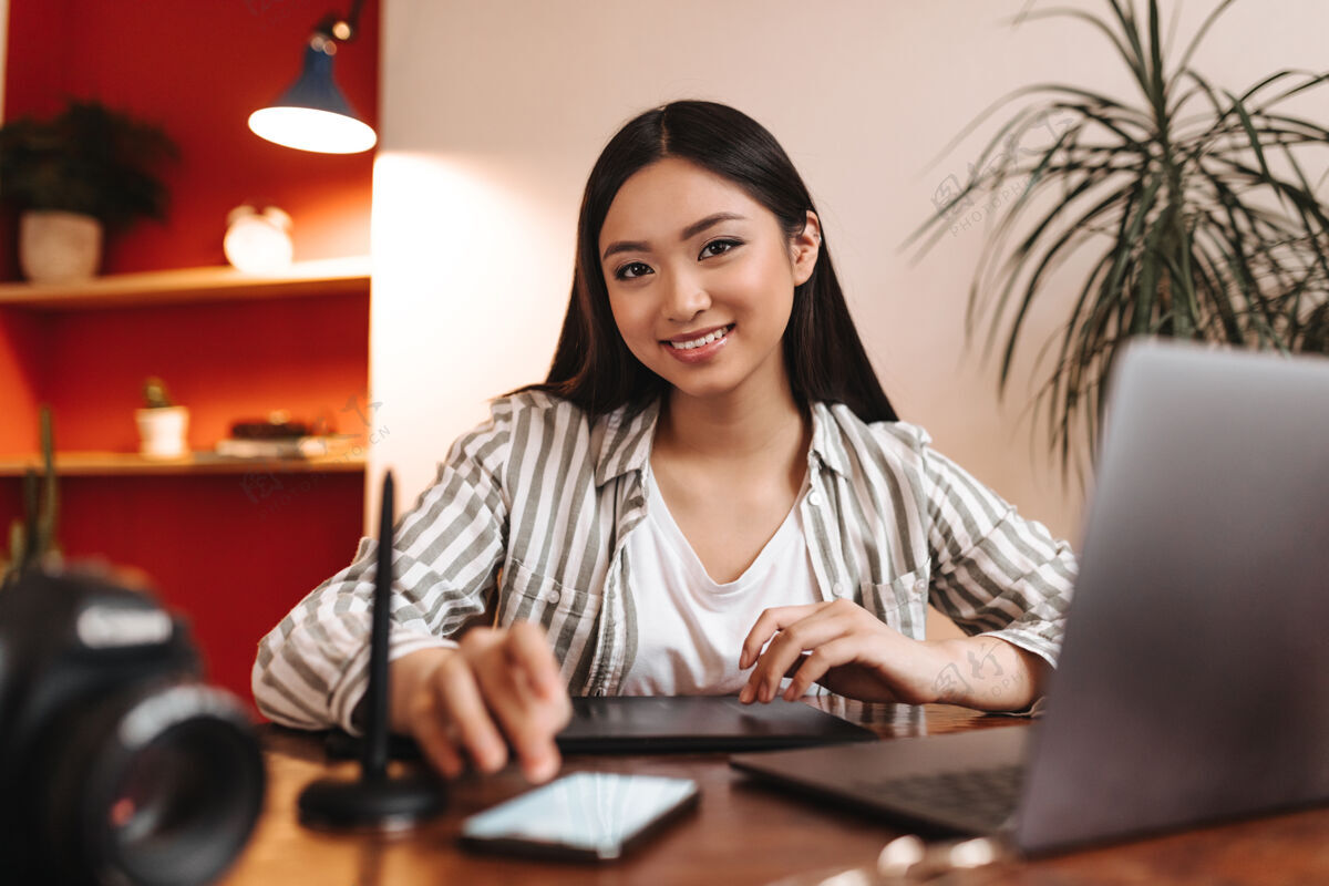笔记穿着条纹衬衫的棕色眼睛女人微笑着 拿着智能手机和笔记本在工作场所摆姿势在线学习漂亮