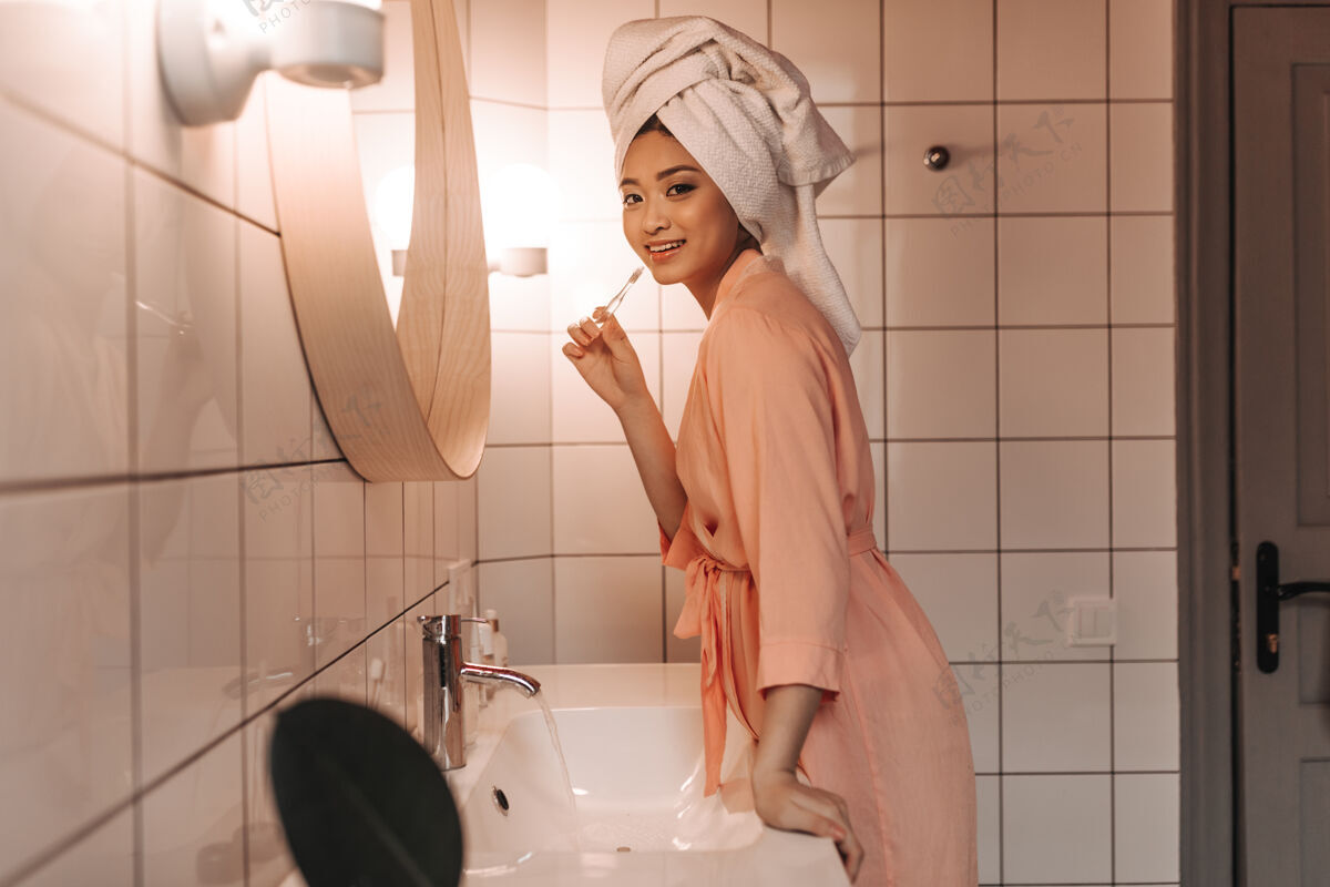 离开穿着粉色长袍的可爱女人刷牙 靠着浴室镜子的墙壁朝前看常规头部化妆