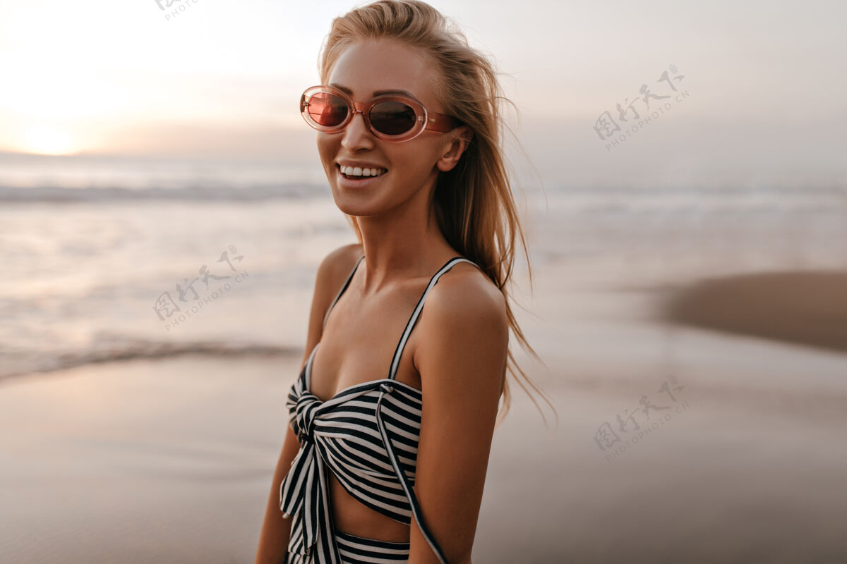 春天美丽快乐的金发女郎 戴着时髦的粉色太阳镜 穿着不规则的条纹上衣 在日落时分微笑着走在沙滩上时尚发髻人
