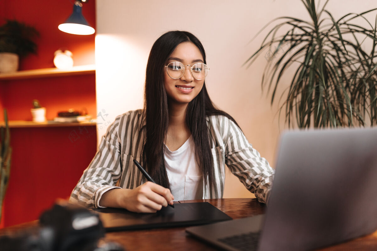 学生身着条纹服装 戴眼镜 面带微笑的女员工看着笔记本电脑 手里拿着笔咖啡快乐室内