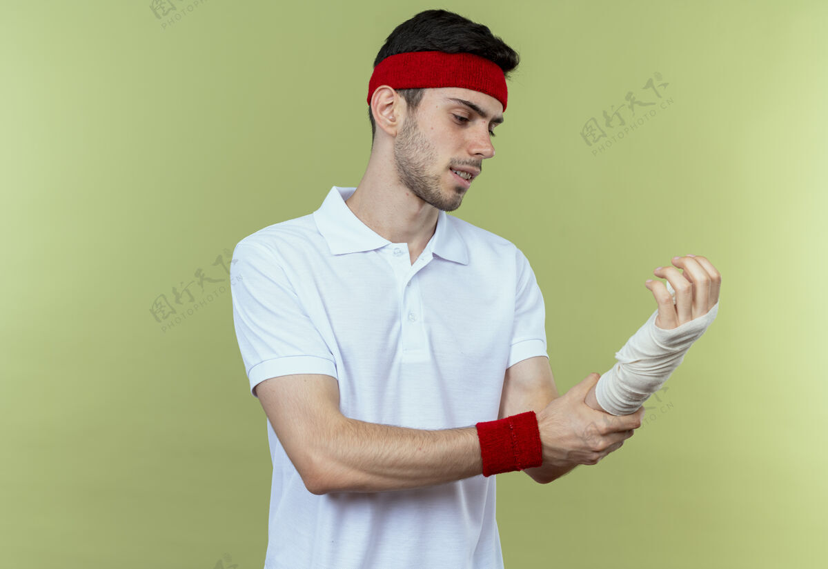 头带戴着头巾的年轻运动型男子站在绿色背景上触摸着他缠着绷带的手腕感到疼痛触摸运动感觉