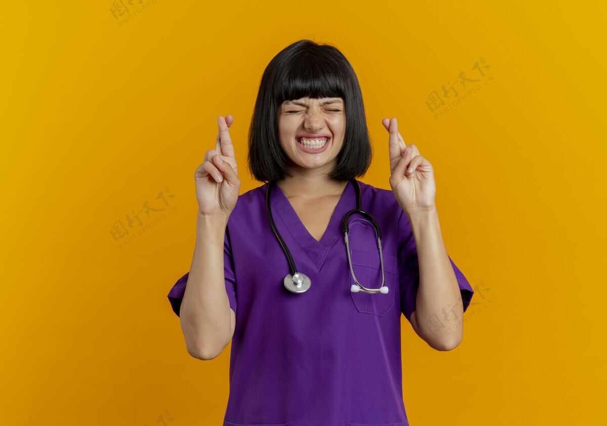 年轻焦急的年轻黑发女医生 穿着制服 手持听诊器 交叉手指 闭着眼睛 隔离在橙色背景上 留有复印空间橙色女性复制