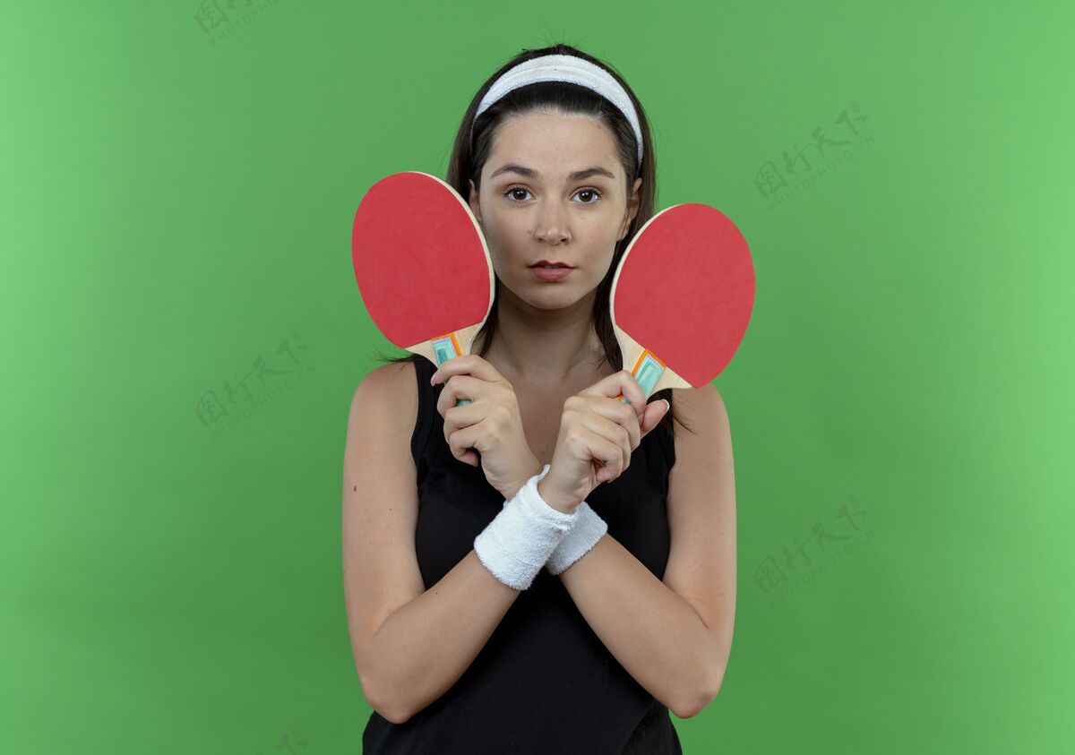 举行戴着头巾拿着球拍打乒乓球的年轻健身女正脸交叉双手站在绿色背景下看着摄像机相机头带立场