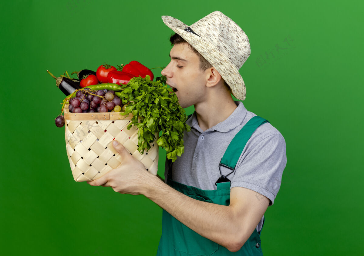 园艺高兴的年轻男园丁戴着园艺帽子拿着和嗅蔬菜篮子隔离在绿色背景与复制空间复制空间嗅