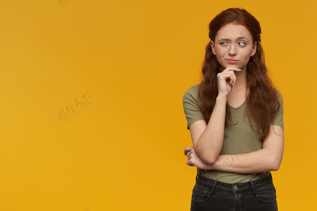 女性好奇 思考的女人 长着姜黄色的头发穿着绿色t恤情感概念触摸她的下巴 抬起眉毛看着左边的复制空间 隔离在橙色的墙上好奇思考欧洲人