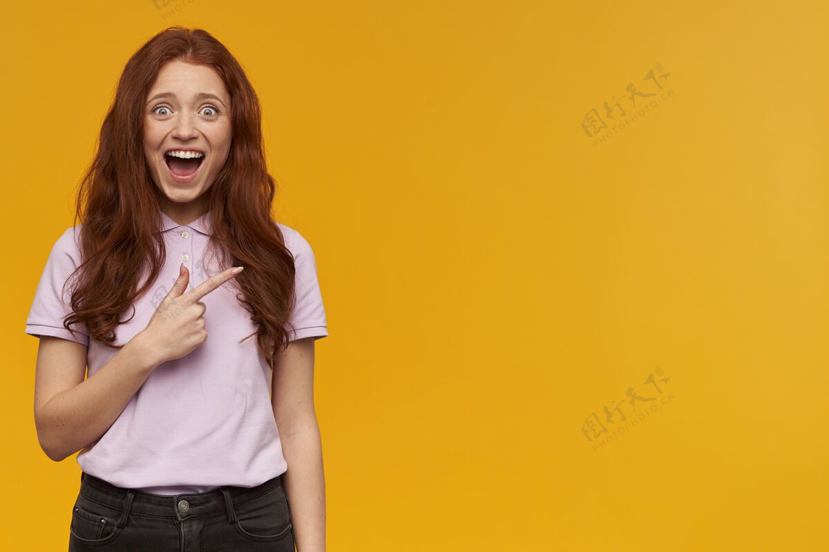 成人兴奋 积极向上的女人 长着一头姜黄色的头发穿着粉色t恤人和情感的概念指着右边的复制空间 隔着橙色的墙手指姜年轻