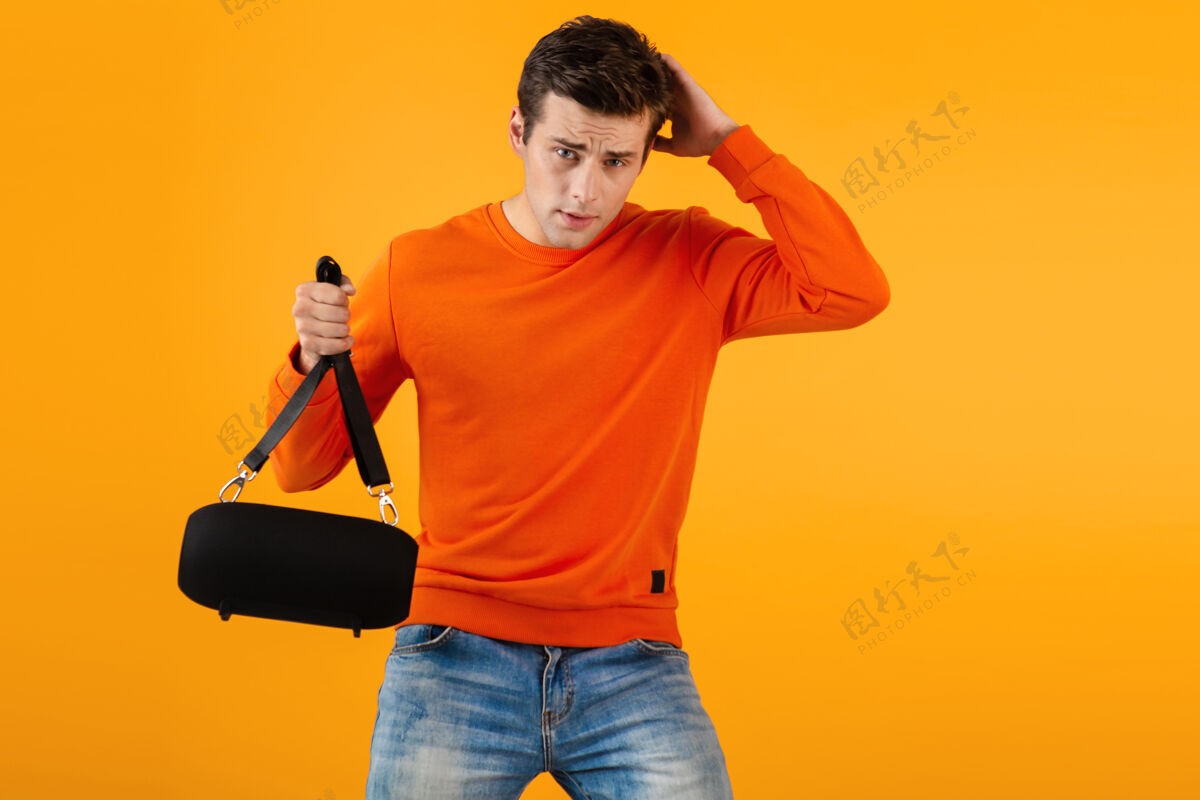 男性时尚微笑的年轻人穿着橙色毛衣手持无线扬声器快乐地听音乐情感时髦快乐