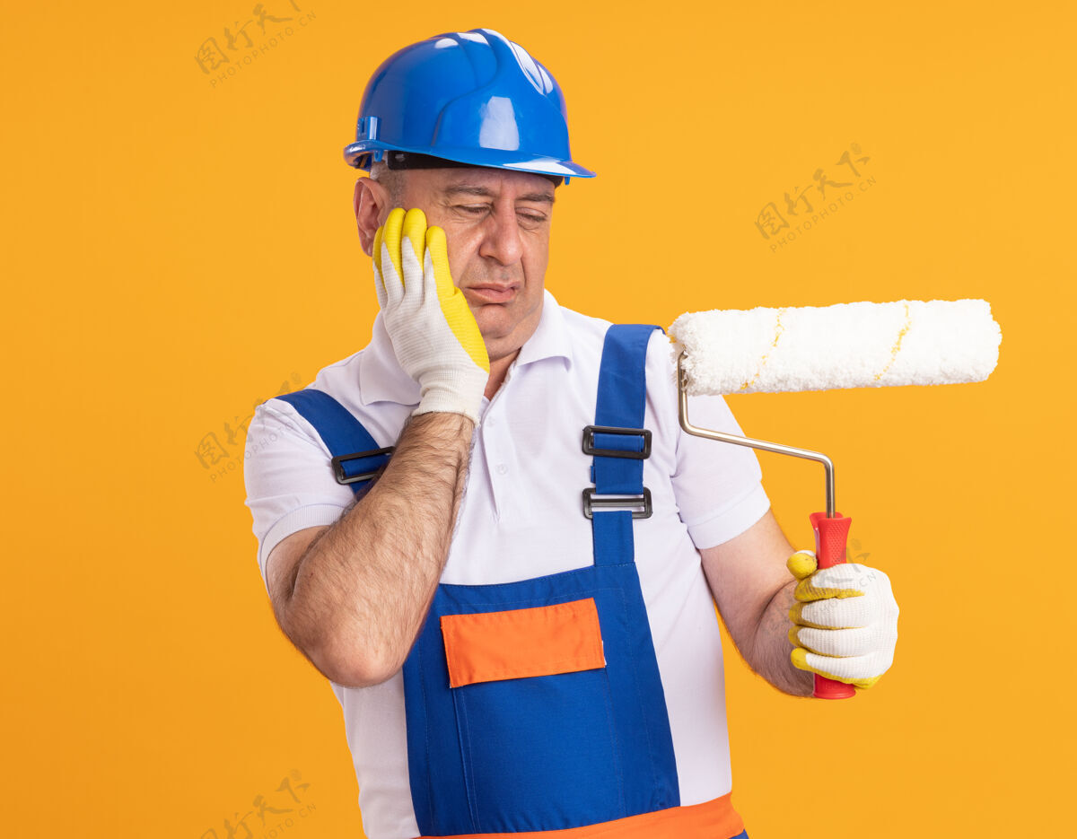 穿着穿着制服的未出租的成年建筑工人戴着防护手套 把手放在脸上 看着隔离在橙色墙上的滚筒刷手套滚轴手