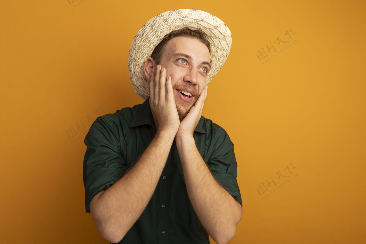 感觉兴奋的金发帅哥 戴着沙滩帽 把手放在脸上 看着橙色墙上孤立的一面人公民英俊