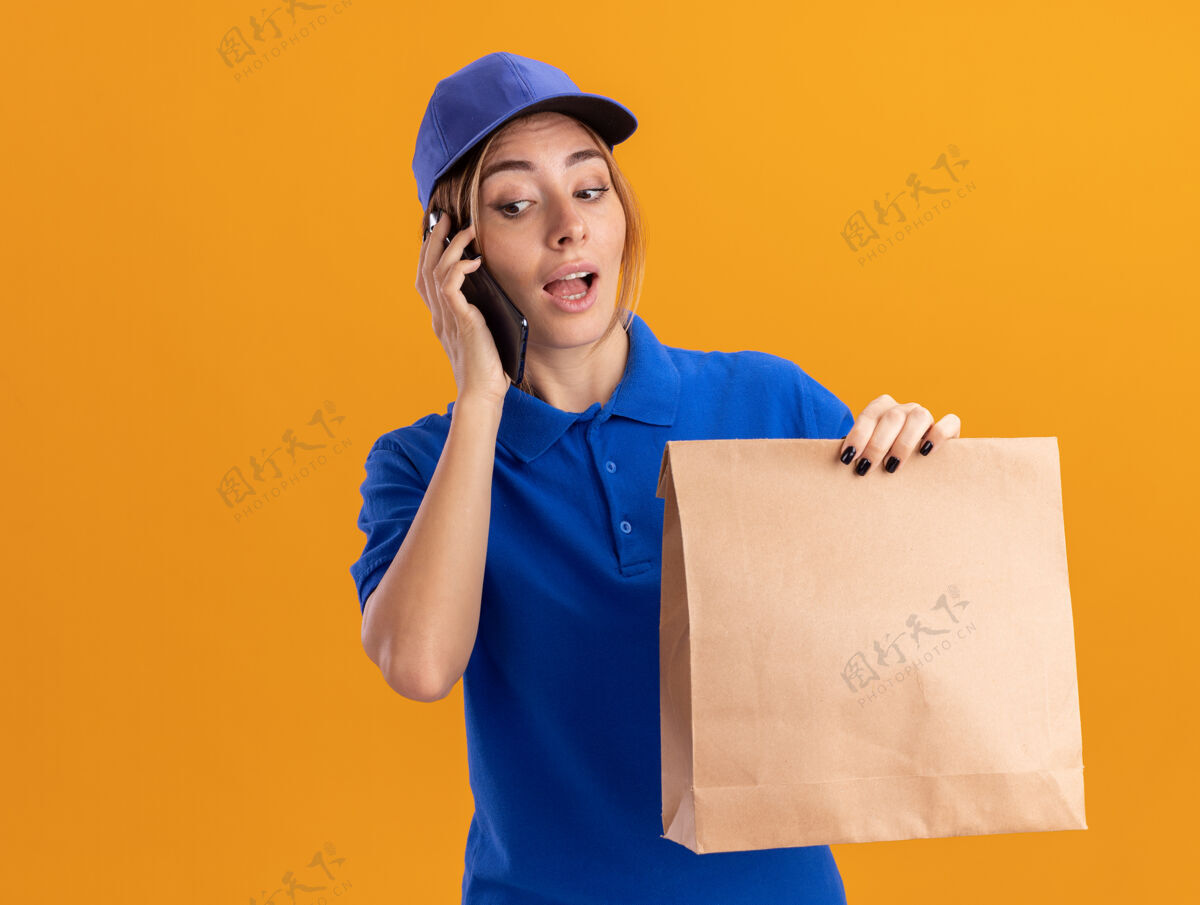 送货一个穿着制服的年轻漂亮的女送货员拿着纸包 隔着橘色的墙上讲着电话人市民制服