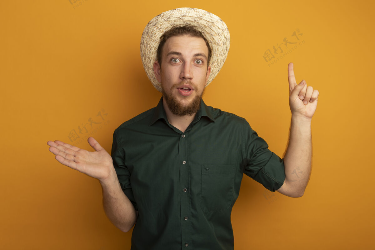 打开一个戴着沙滩帽的金发帅哥惊讶地张开手 孤立地指着橙色的墙上站帅哥手