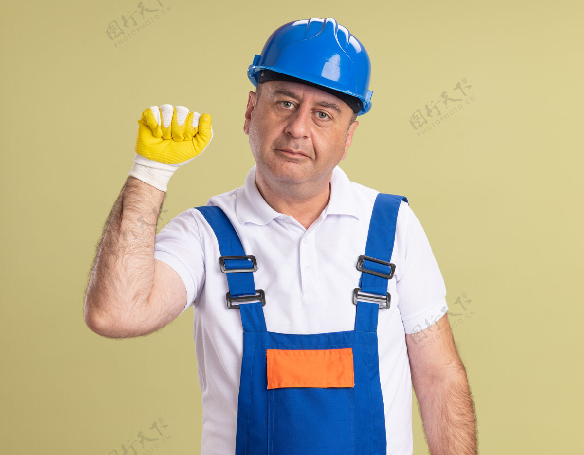 感情高兴的成年建筑工人穿着制服 戴着防护手套 保持拳头在橄榄绿的墙上孤立橄榄保护立场