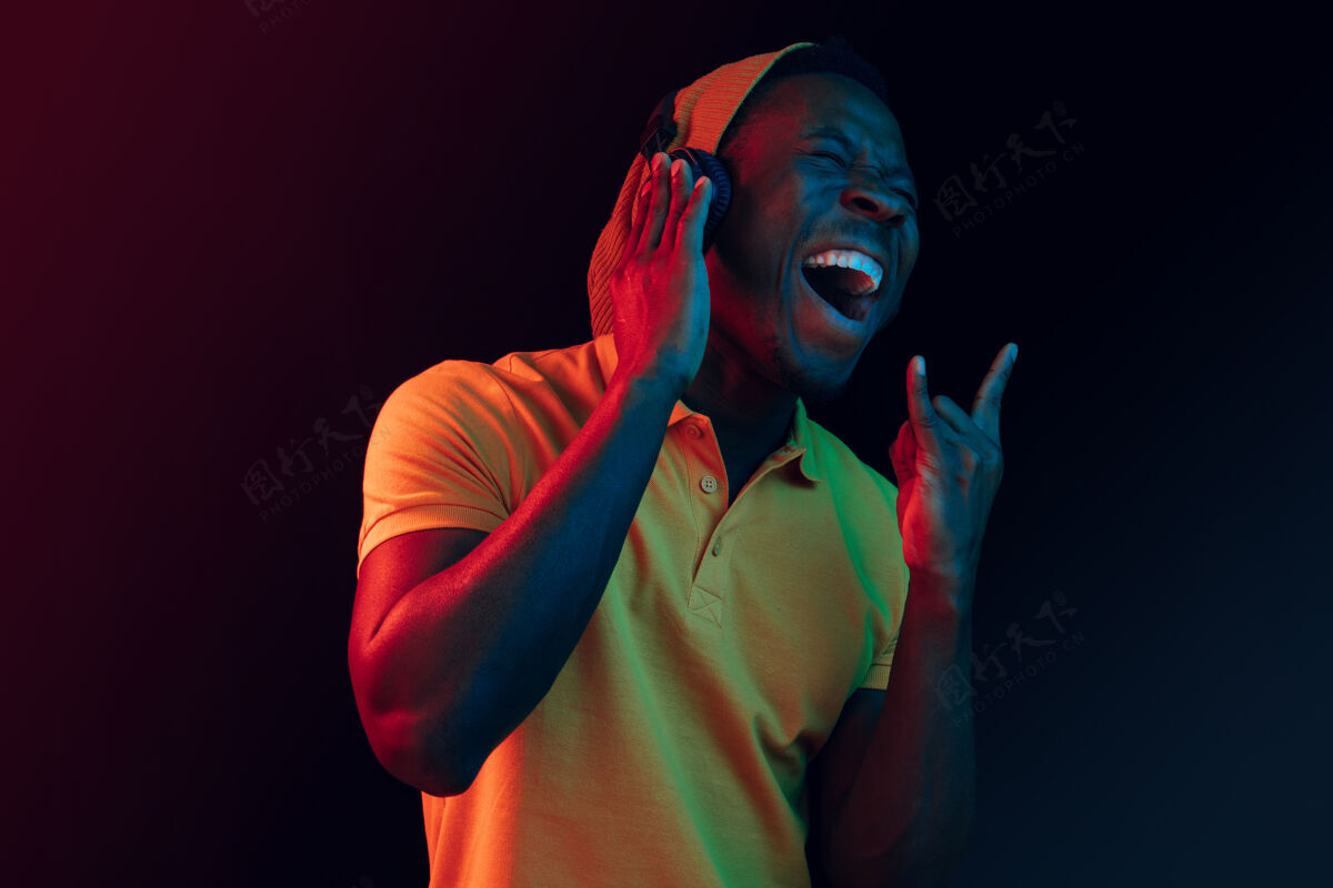 非洲年轻帅气快乐的时髦男人在霓虹灯下用耳机在黑工作室听音乐迪斯科舞厅 夜总会 嘻哈风格 积极的情绪 面部表情 舞蹈概念音频脸夜总会