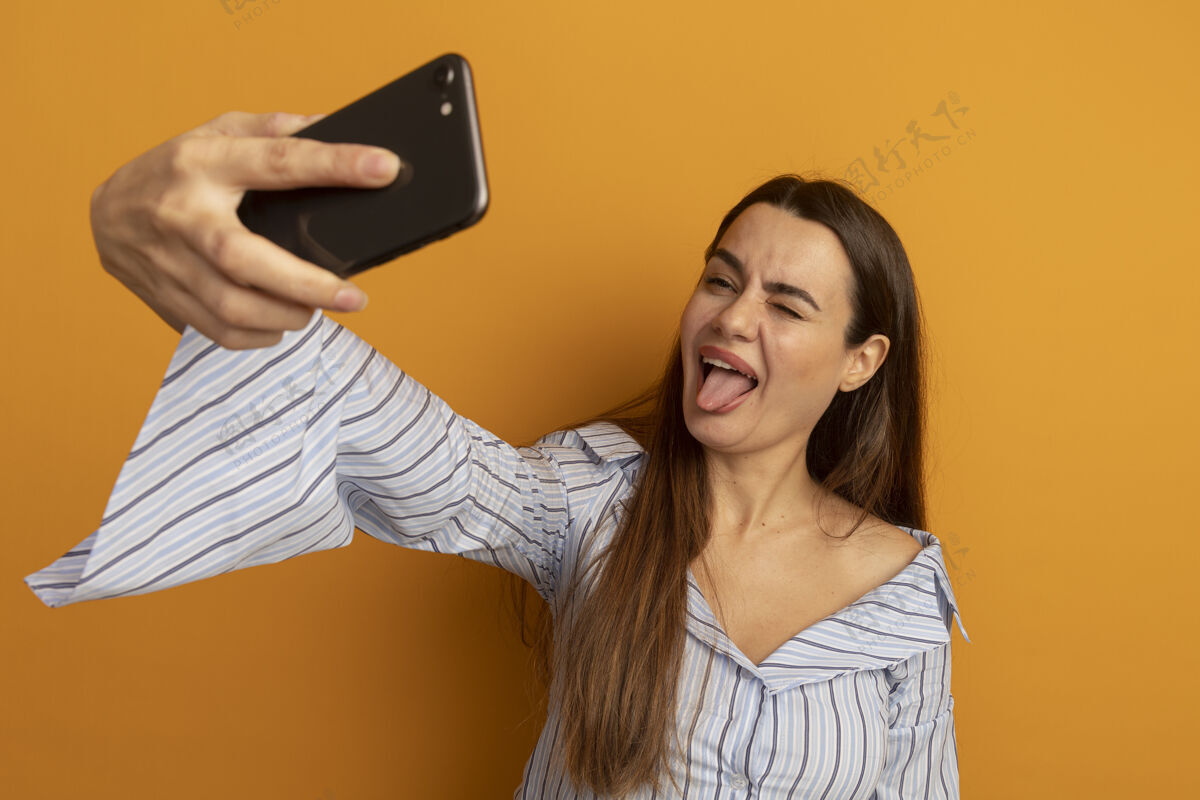 拿着有趣的漂亮女人眨着眼睛结结巴巴地伸出舌头拿着手机看自拍隔离在橙色的墙上姿势舌头搞笑