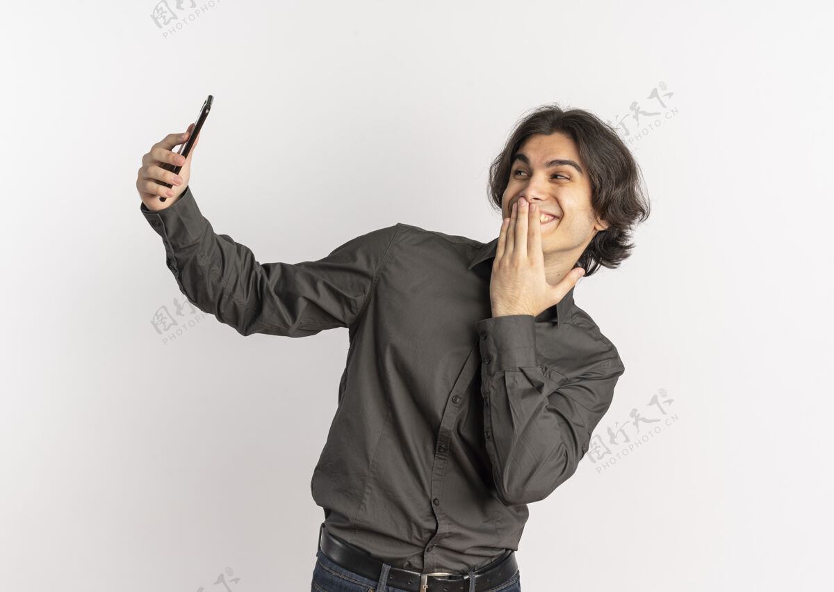 年轻年轻快乐英俊的白人男子把手放在嘴上 看着手机 手机被隔离在白色背景上 留有复印空间英俊嘴手