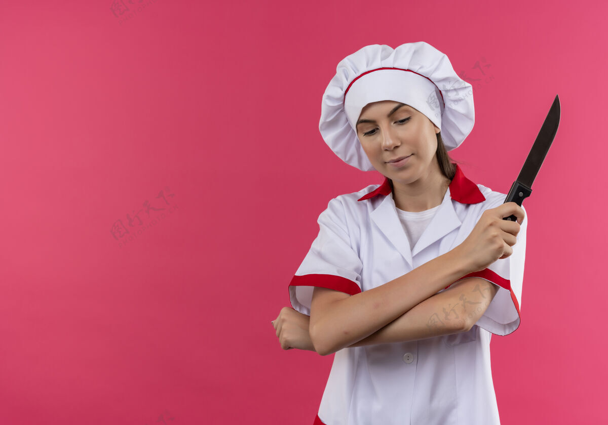 女孩身着厨师制服的年轻快乐的白人厨师女孩交叉双臂 手持隔离在粉色背景上的刀 并留有复印空间交叉厨师手臂