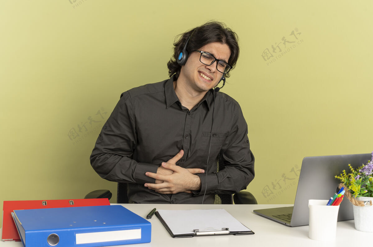 使用年轻烦躁的上班族戴着耳机戴着眼镜坐在办公桌旁 拿着办公工具用笔记本电脑把手放在肚子上 隔离在绿色背景上 留有复印空间笔记本电脑坐着办公桌
