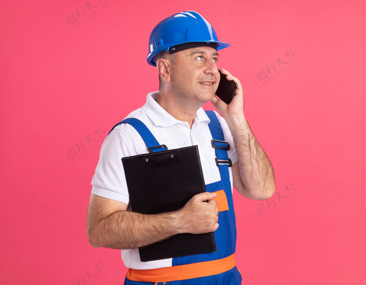人穿着制服的成年建筑工人高兴地拿着剪贴板 一边打电话 一边看着粉红色墙上孤立的一面成人公民姿势