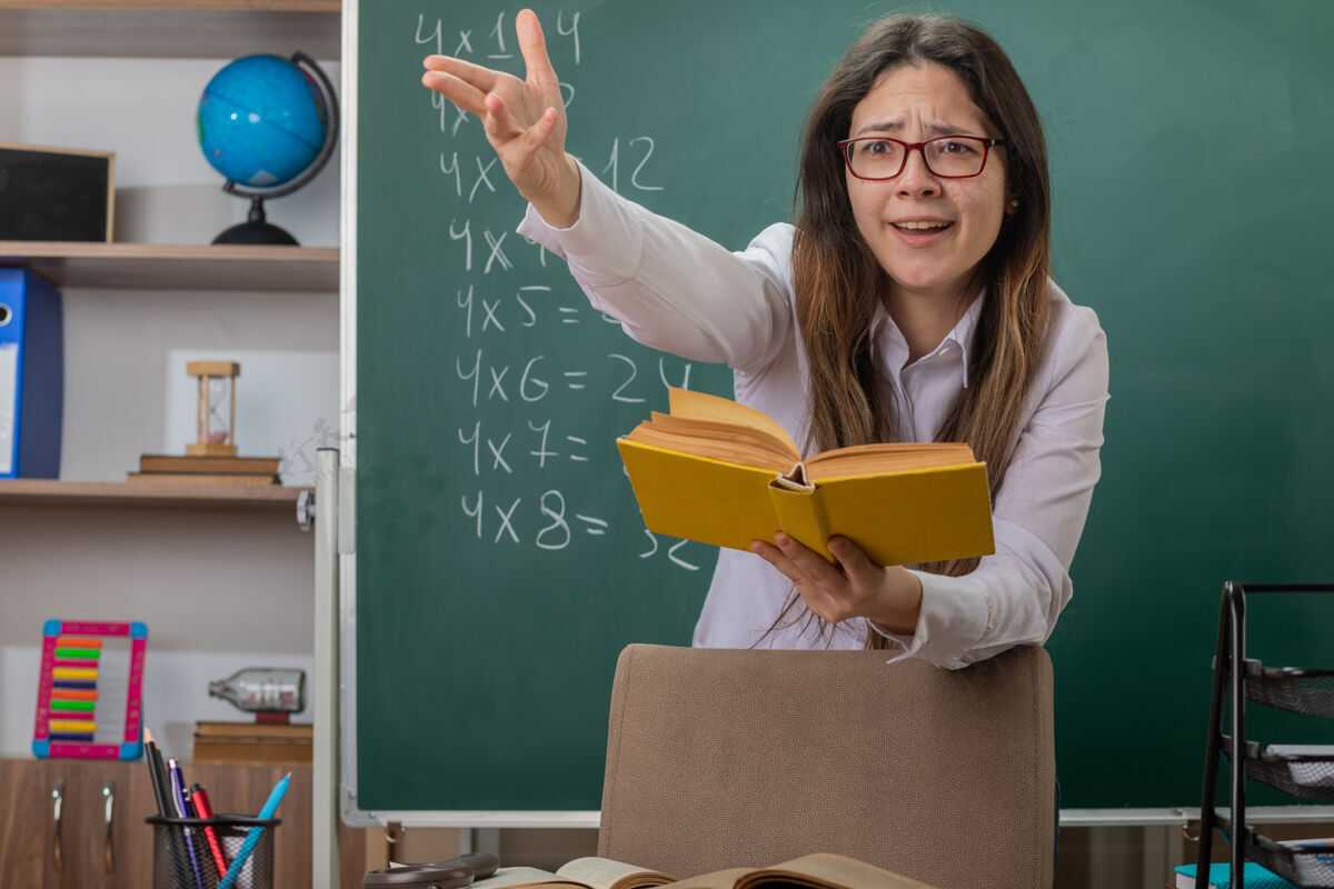 拿着戴眼镜的年轻女教师站在教室的黑板前 手捧讲解课的书 感到困惑和不快女人不高兴老师