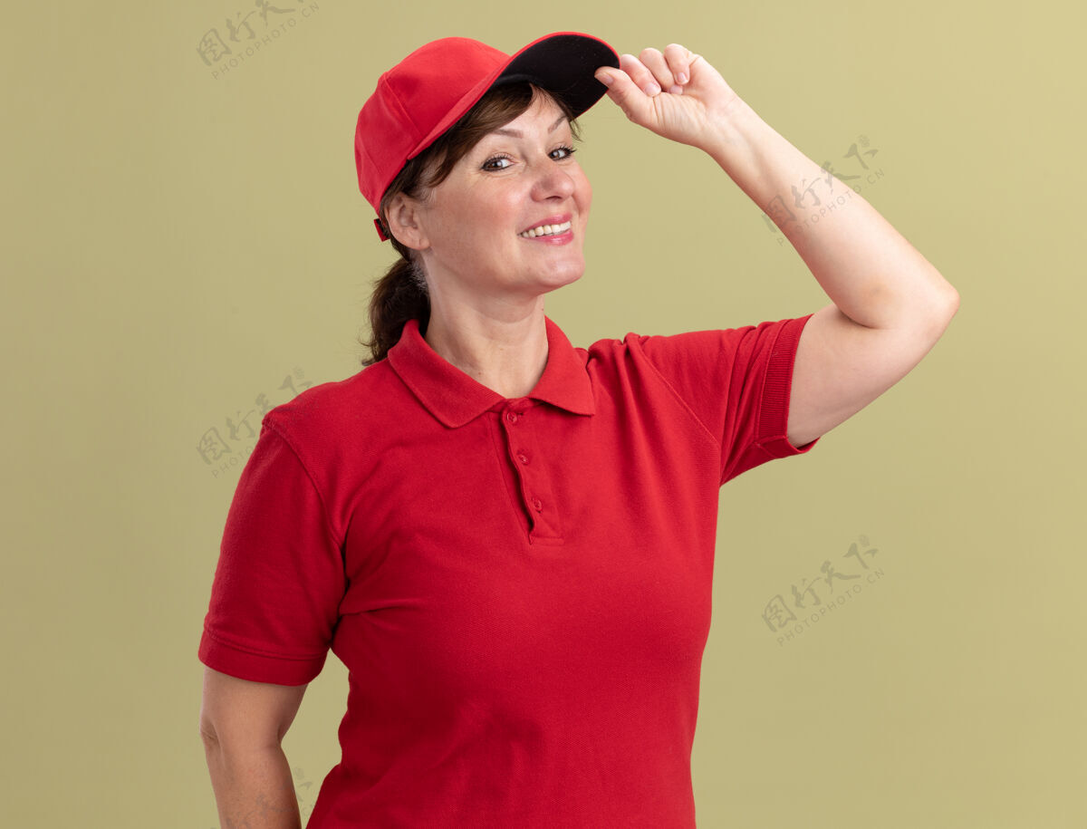 触摸身穿红色制服 头戴帽子的中年女送货员站在绿色的墙上 微笑着自信地抚摸着她的帽子女人微笑帽子
