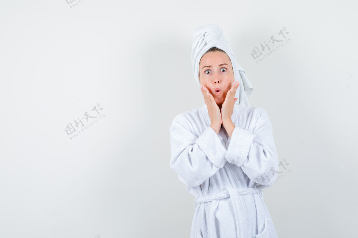 年轻照片中的年轻女子手放在脸颊上 穿着白色浴衣 拿着毛巾 脸上露出惊讶的表情时尚成人皮肤
