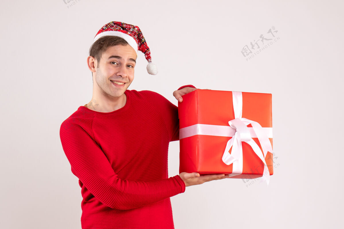 快乐正面图：戴着圣诞帽的年轻人拿着白色背景的红色礼盒漂亮礼物举行