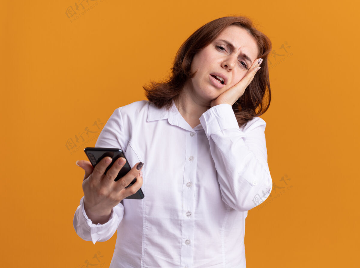 困惑身穿白衬衫 手持智能手机的年轻女子站在橙色的墙上 困惑而不安地看着前方衬衫年轻女人
