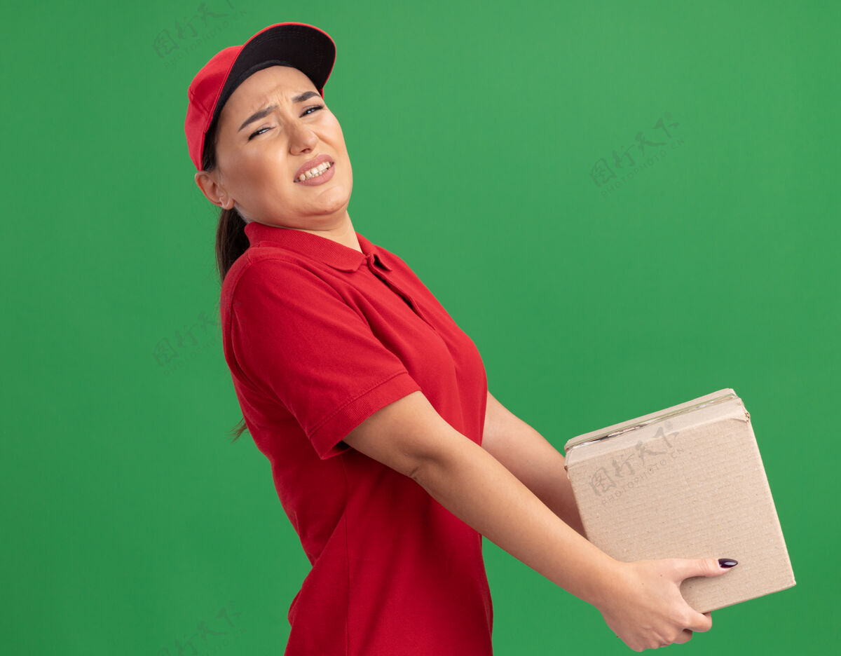 纸板年轻的送货员身穿红色制服 戴着帽子 手里拿着沉重的纸板箱 站在绿色的墙上 神情疲惫 神情烦躁表情帽子站着