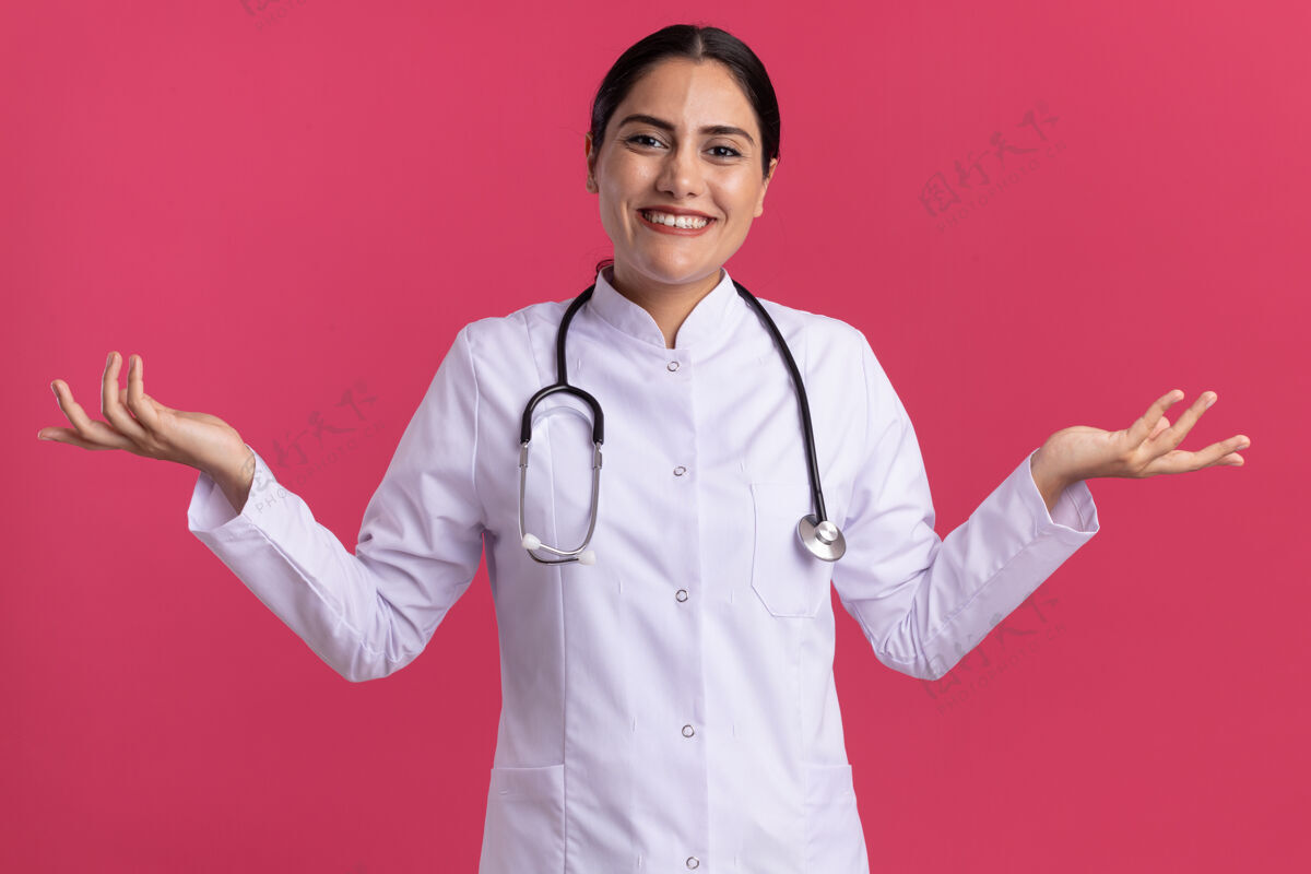 举起来年轻的女医生 身穿医用外套 手持听诊器 面带微笑 双臂高举 站在粉红色的墙上外套女人微笑