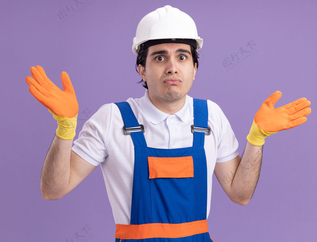 手套年轻的建筑工人穿着建筑制服 戴着橡胶手套戴着安全帽 站在紫色的墙上 困惑而不确定地看着前方头盔困惑年轻