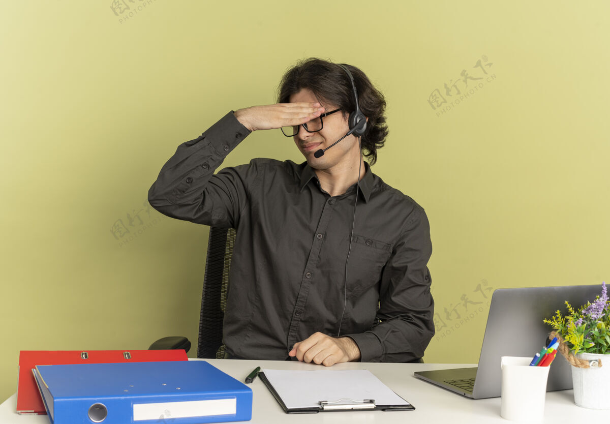 光学年轻恼怒的上班族戴着耳机戴着眼镜坐在办公桌旁 拿着办公工具用笔记本电脑 把头隔离在绿色背景上 留着复印空间坐着耳机拿着