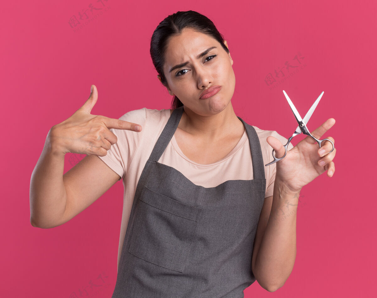 拿着年轻漂亮的女理发师站在粉红色的墙上 手里拿着剪刀 用食指指着它 看起来很自信手指指着女人